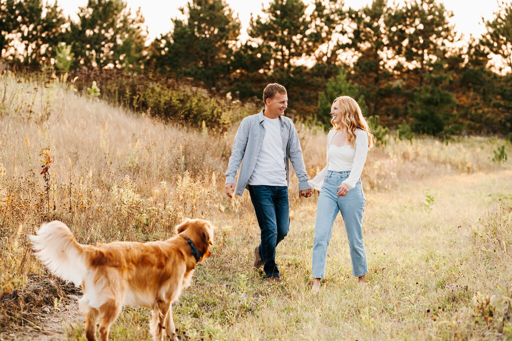 Engaged couple sharing a playful moment with their golden retriever in a lush Bemidji field during golden hour, embodying the romance of nature