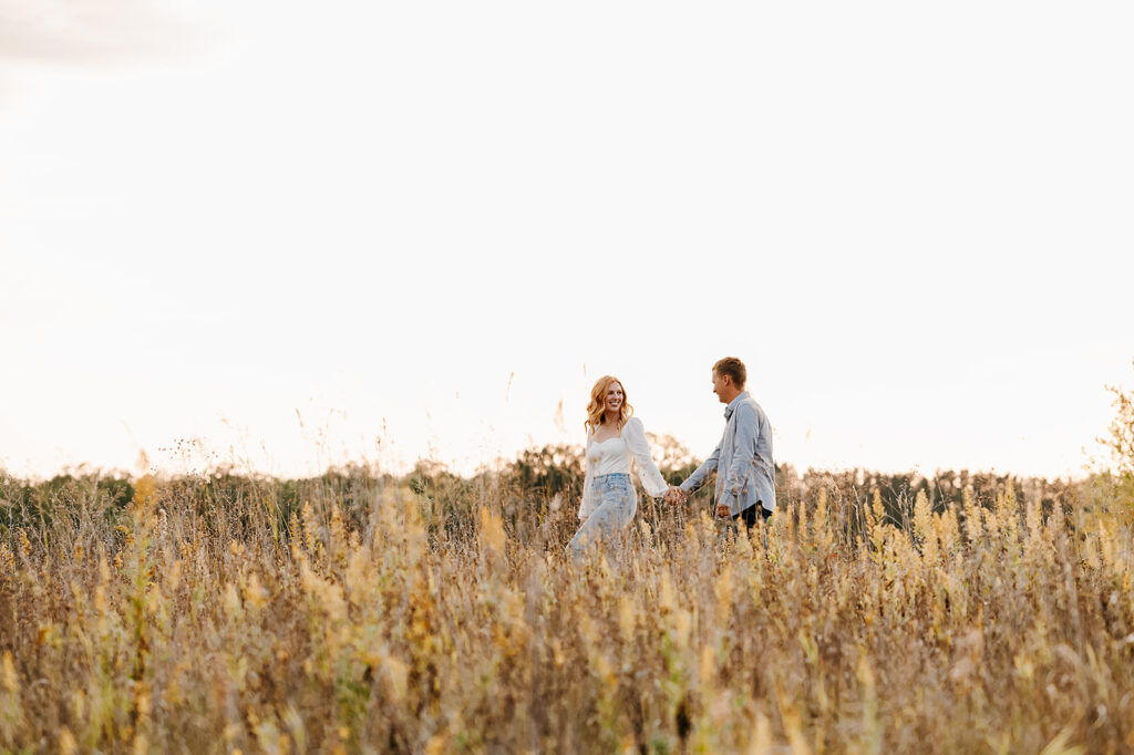 Future bride and groom walking hand in hand through a scenic field in Bemidji, with the warm golden hour sunlight enhancing the romantic atmosphere