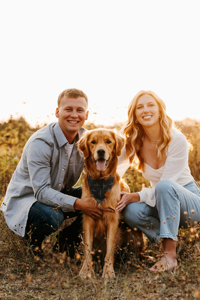 Engaged couple sharing a playful moment with their golden retriever in a lush Bemidji field during golden hour