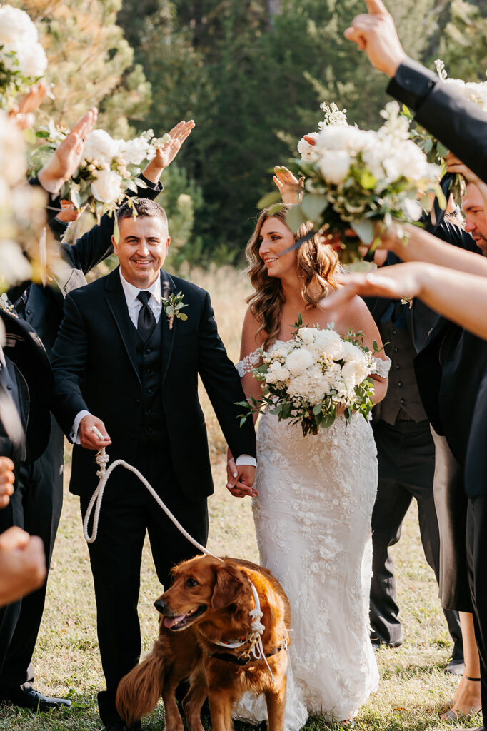 bride and groom and their golden retriever walking through a hand bridge of their wedding party