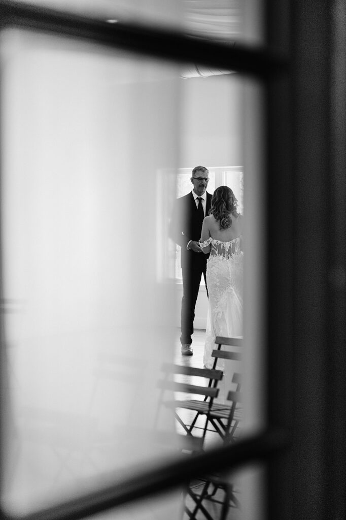 Long distance, black and white photos capturing bride and dad's first look reactions through a window, seeing their happy tears and so much love