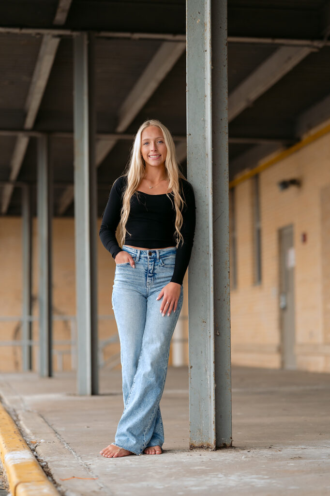 High school senior posing next to a rustic building in a casual attire of black top and jeans