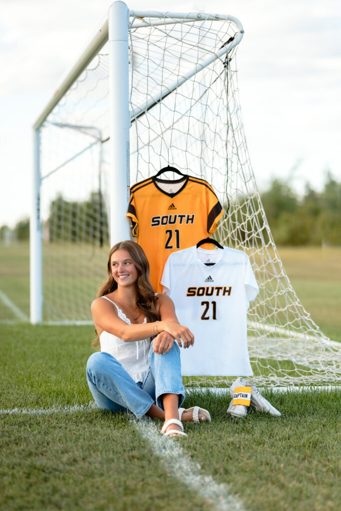 senior photos in minnesota featuring a girl sitting in a football field next to the goal with jerseys hanging in the background