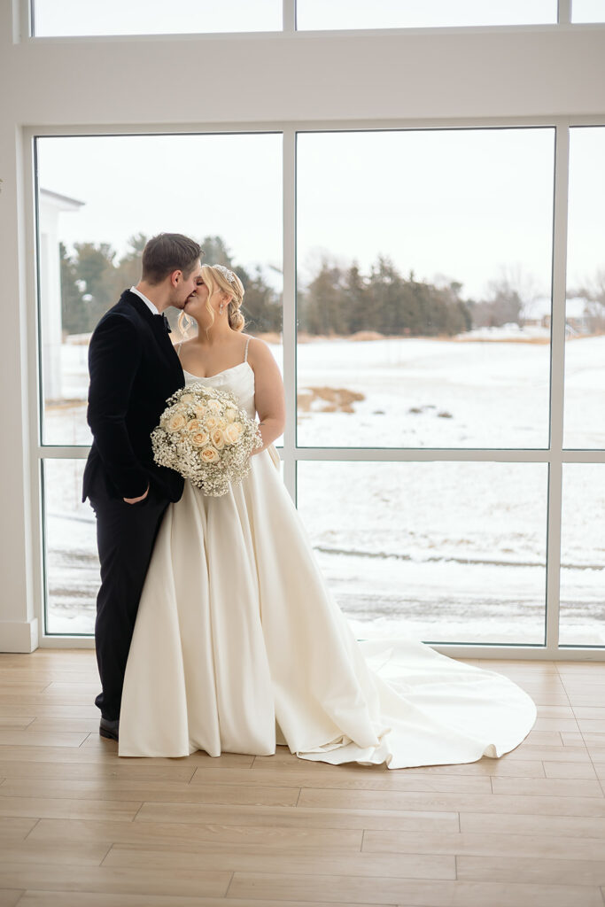 Bride and groom kissing in front of the large windows at The Aisling during winter wedding