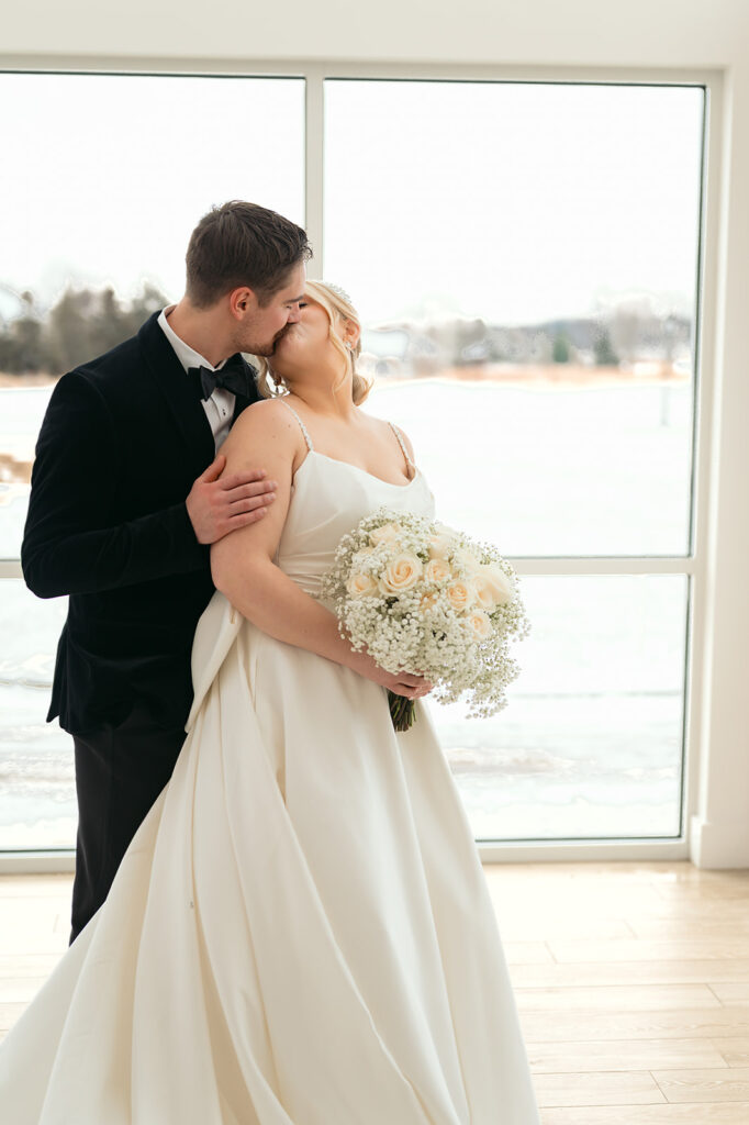 Bride and groom kissing in front of the large windows at The Aisling during winter wedding