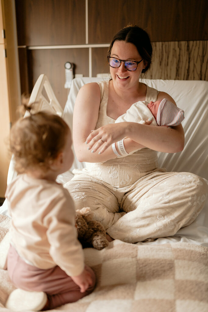 mom holding a newborn in the hospital as they introduce toddler to the baby