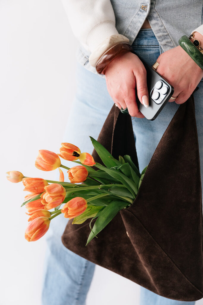Smiling business owner holding her phone, tote bag and orange tulips during a personal branding photoshoot.