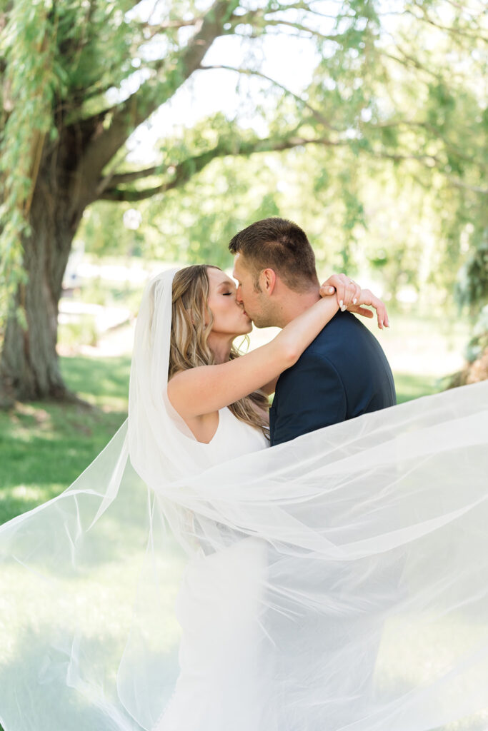 Bride and groom sharing a kiss beneath a willow tree as the bride’s veil flows around them